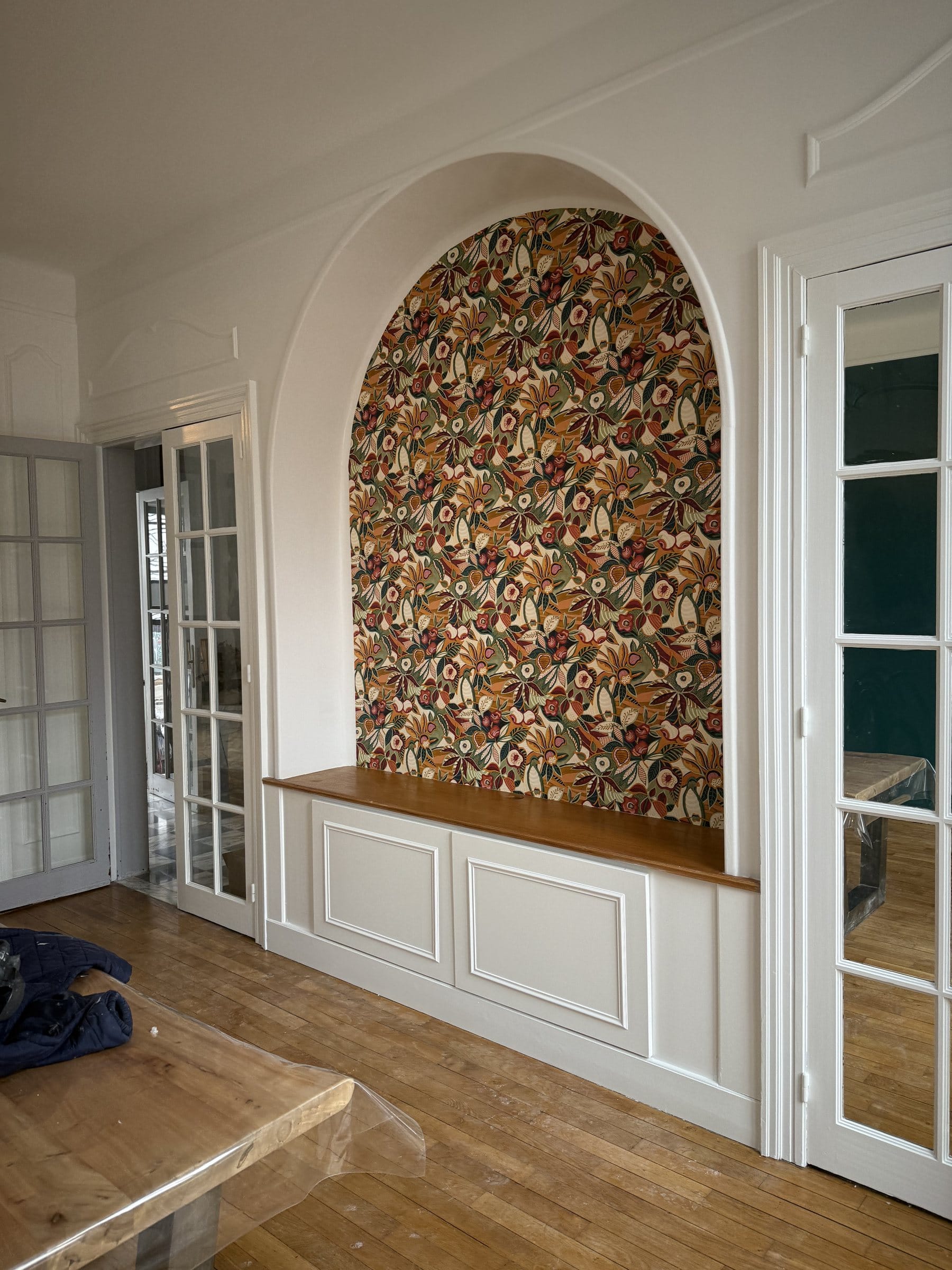 Niche en arc tapissée d’un papier peint floral, banc et boiseries blancs, portes vitrées